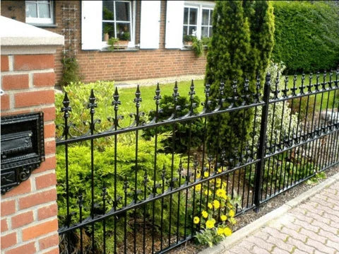 Sort smedejernshegn foran et hus med have og en murstenspille med postkasse. Gulde blomster ved flisegangen.
